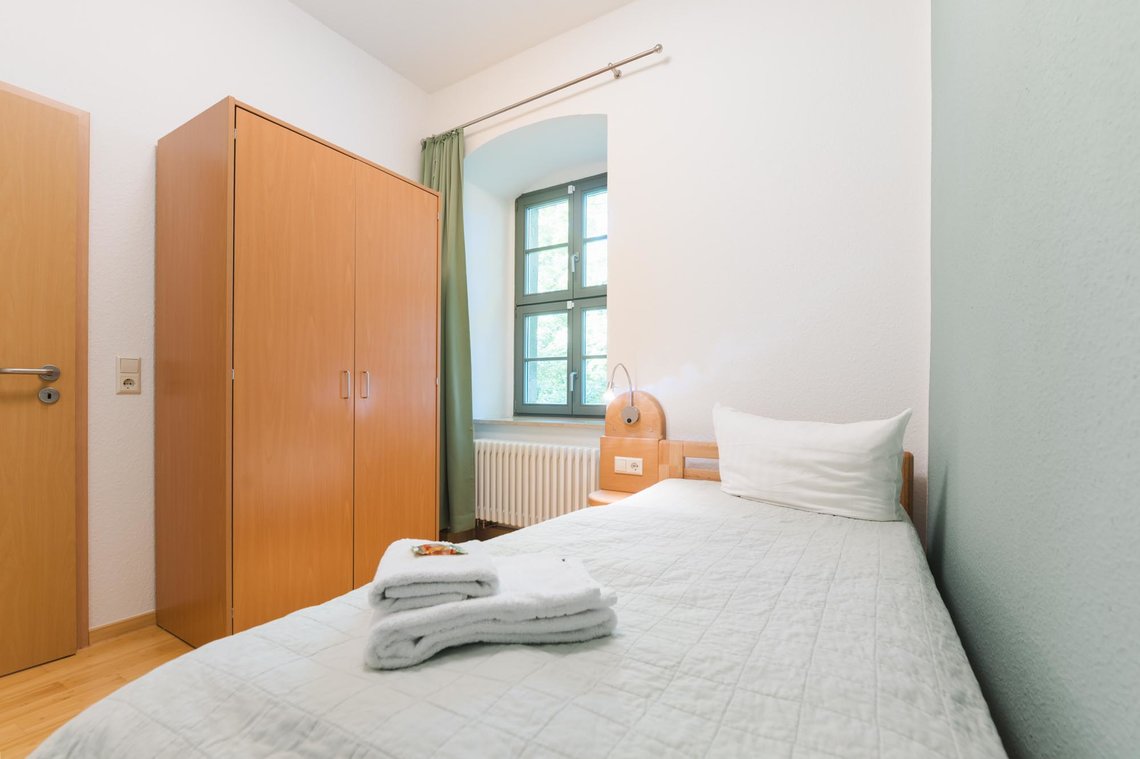 Ein kleines Zimmer mit einem Fenster,  einem Bett mit 2 Handtüchern, einem Schrank und einer Tür.