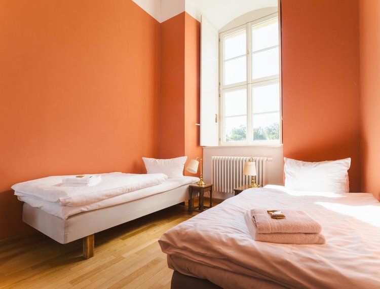 Helles Gästezimmer mit zwei Einzelbetten, warmen orangefarbenen Wänden und großem Fenster. Auf den Betten liegen gefaltete Handtücher, daneben stehen kleine Nachttische mit Lampen.