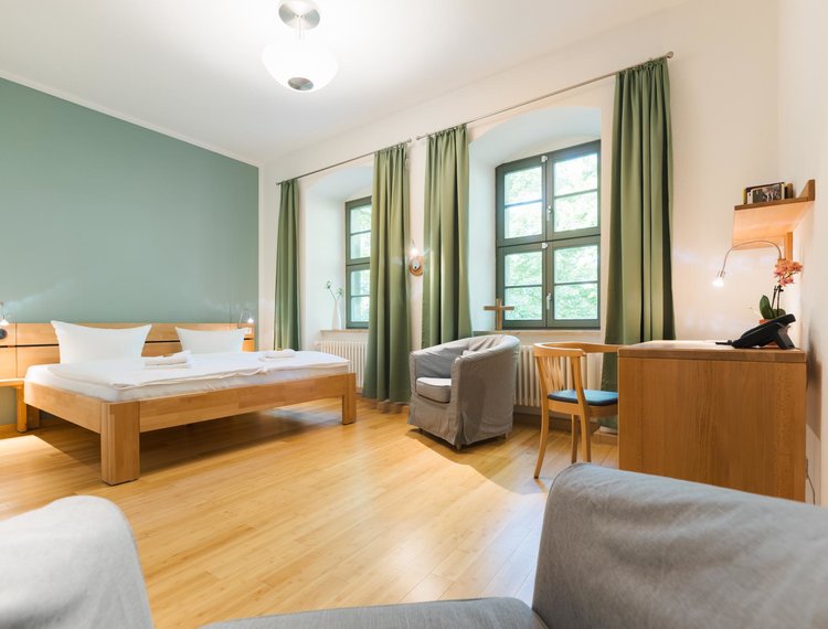Aus der Sicht eines grauen Sessels sieht man ein Hotelzimmer mit einer weißen und einer grünen Wand und 2 Fenstern mit grünen Vorhängen. Links vor der grünen Wand steht ein Doppelbett mit zwei Lampen. Rechts davon steht ein grauer Sessel vor dem Fenster. In dem Fenster steht ein Kreuz. An der rechten Wand steht ein Schreibtisch mit einem Stuhl. Auf dem Schreibtisch steht eine Blume und ein Telefon. Darüber hängt ein Bücherregal.