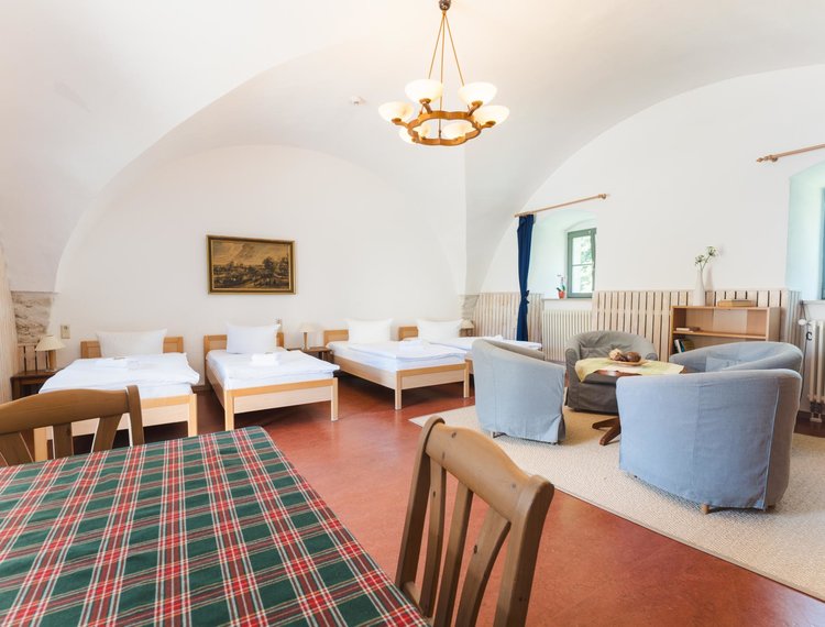 Helles, geräumiges Zimmer mit zwei Einzelbetten links und mit zwei Einzelbetten rechts, einem kleinen Sitzbereich mit vier Sesseln und einem runden Tisch rechts sowie einem Esstisch im Vordergrund. An der Wand hängt ein Gemälde, und ein Fenster mit blauem Rahmen lässt Tageslicht herein.