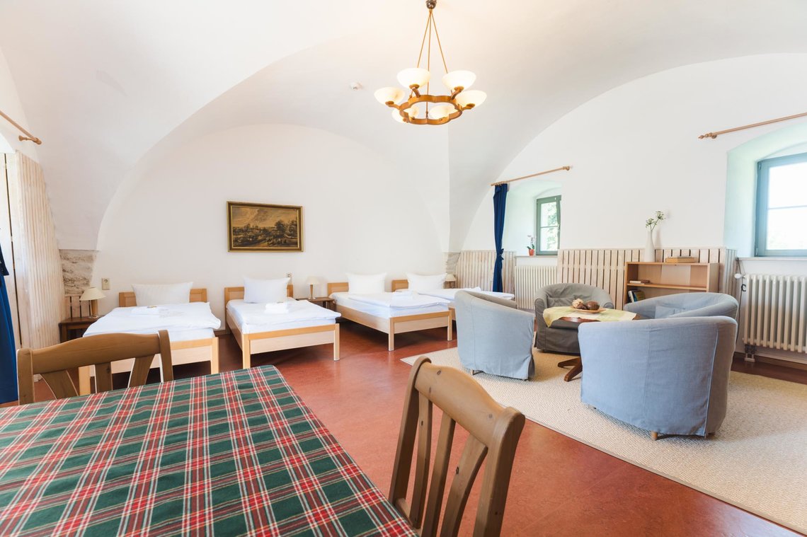 Helles, geräumiges Zimmer mit zwei Einzelbetten links und mit zwei Einzelbetten rechts, einem kleinen Sitzbereich mit vier Sesseln und einem runden Tisch rechts sowie einem Esstisch im Vordergrund. An der Wand hängt ein Gemälde, und ein Fenster mit blauem Rahmen lässt Tageslicht herein.