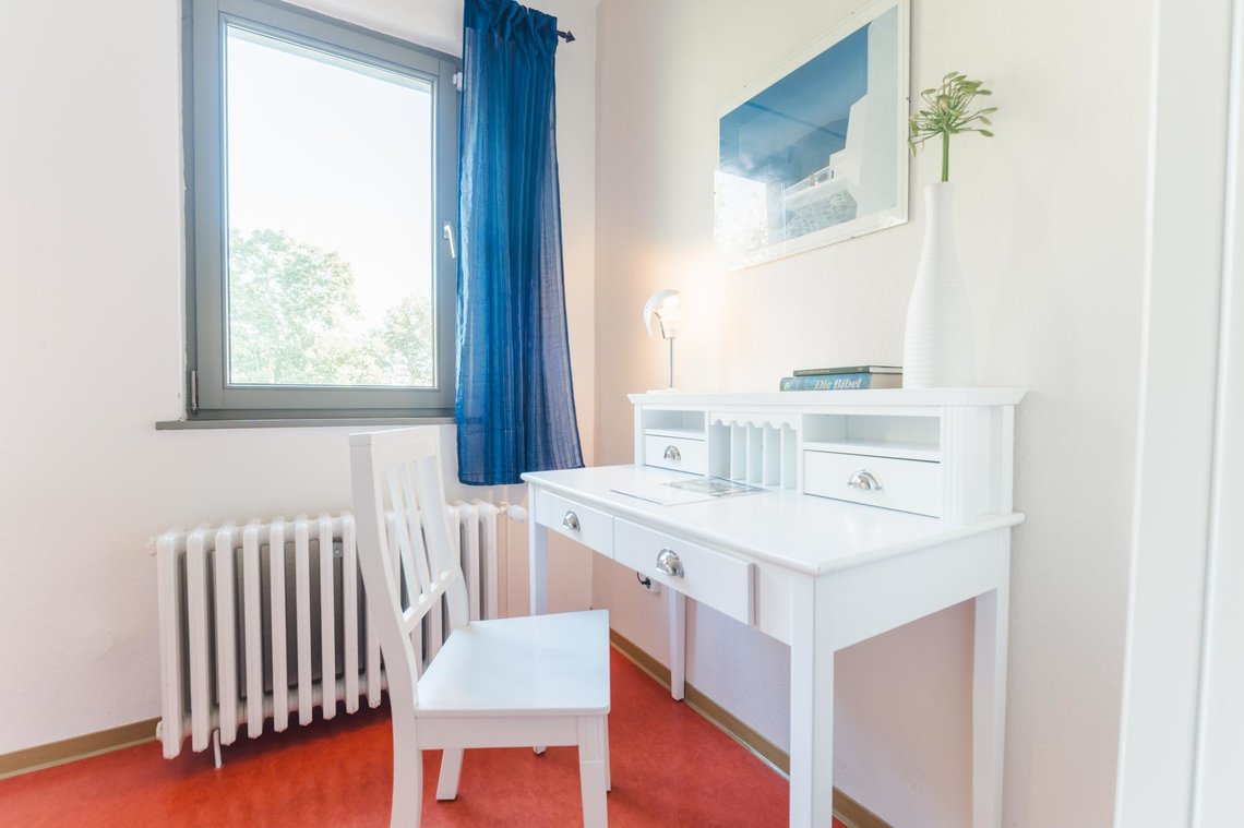 In einem weißen Raum mit rotem Boden steht vor einem Fenster ein weißer Schreibtisch mit Stuhl. Auf dem Schreibtisch steht eine Lampe, eine Blume und eine Bibel. Über dem Schreibtisch hängt ein Bild. An dem Fenster hängt eine blaue Gardine.