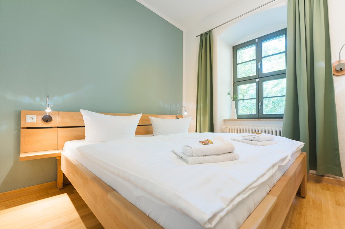 Ein Hotelzimmer mit einer weißen und einer grünen Wand und 2 Fenstern mit grünen Vorhängen. Links vor der grünen Wand steht ein Doppelbett mit zwei Lampen. 