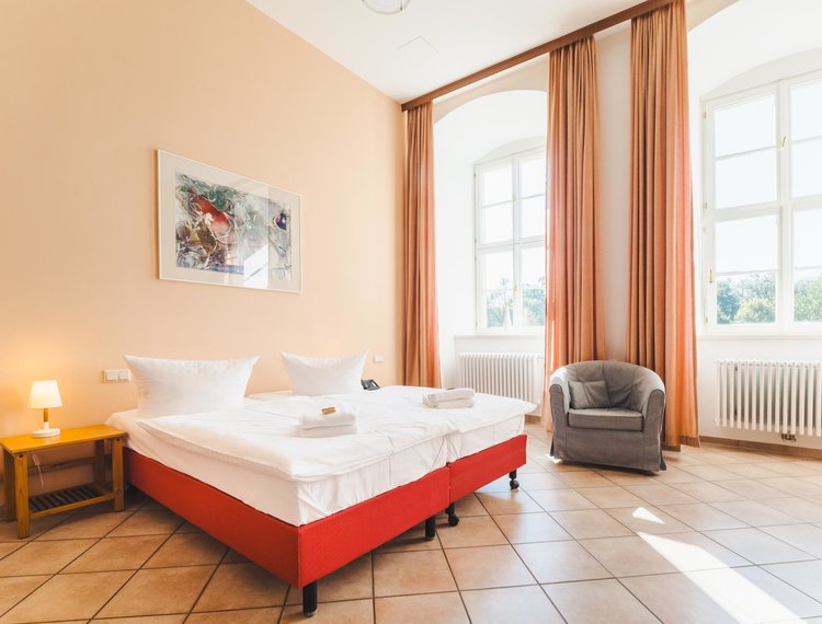 Ein großes Doppelbett und ein Sessel stehen in einem geräumigen Raum mit hohen Fenstern. 