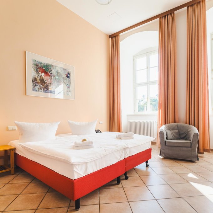 Ein großes Doppelbett und ein Sessel stehen in einem geräumigen Raum mit hohen Fenstern.