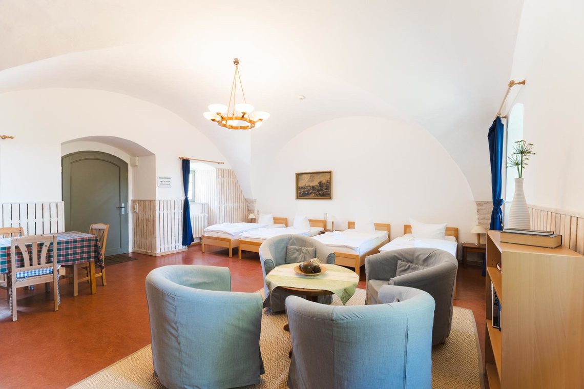 Ein geräumiges, helles Gästezimmer mit gewölbter Decke. Rechts im Raum stehen vier gepolsterte Sessel um einen runden Tisch mit einer Schale. Dahinter befinden sich vier Einzelbetten in einer Reihe. Links steht ein Esstisch mit kariertem Tischtuch. Eine Tür mit Rundbogen führt nach draußen. Ein Fenster mit blauen Vorhängen bringt Tageslicht in den Raum und daneben steht ein Regal mit einer weißen Vase.