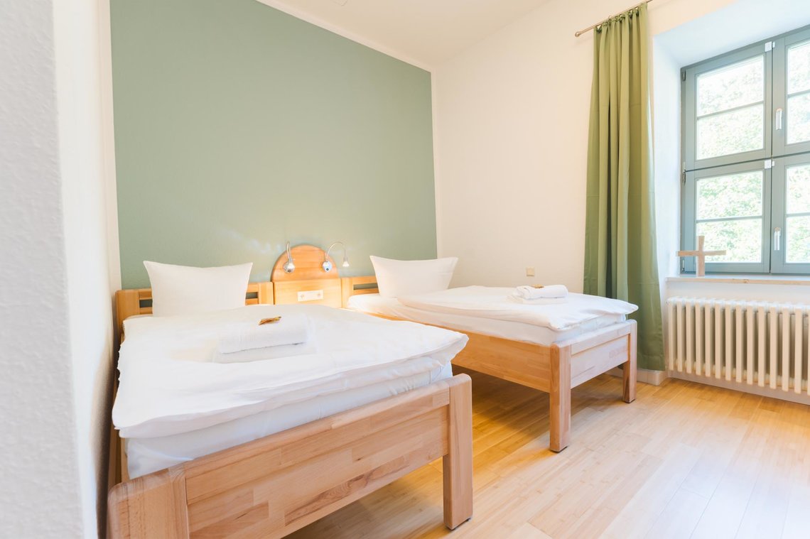 Ein helles weißes Zimmer mit einem Fenster mit grünen Vorhängen. An einer grünen Wand stehen zwei Einzelbetten mit zwei Lampen dazwischen.