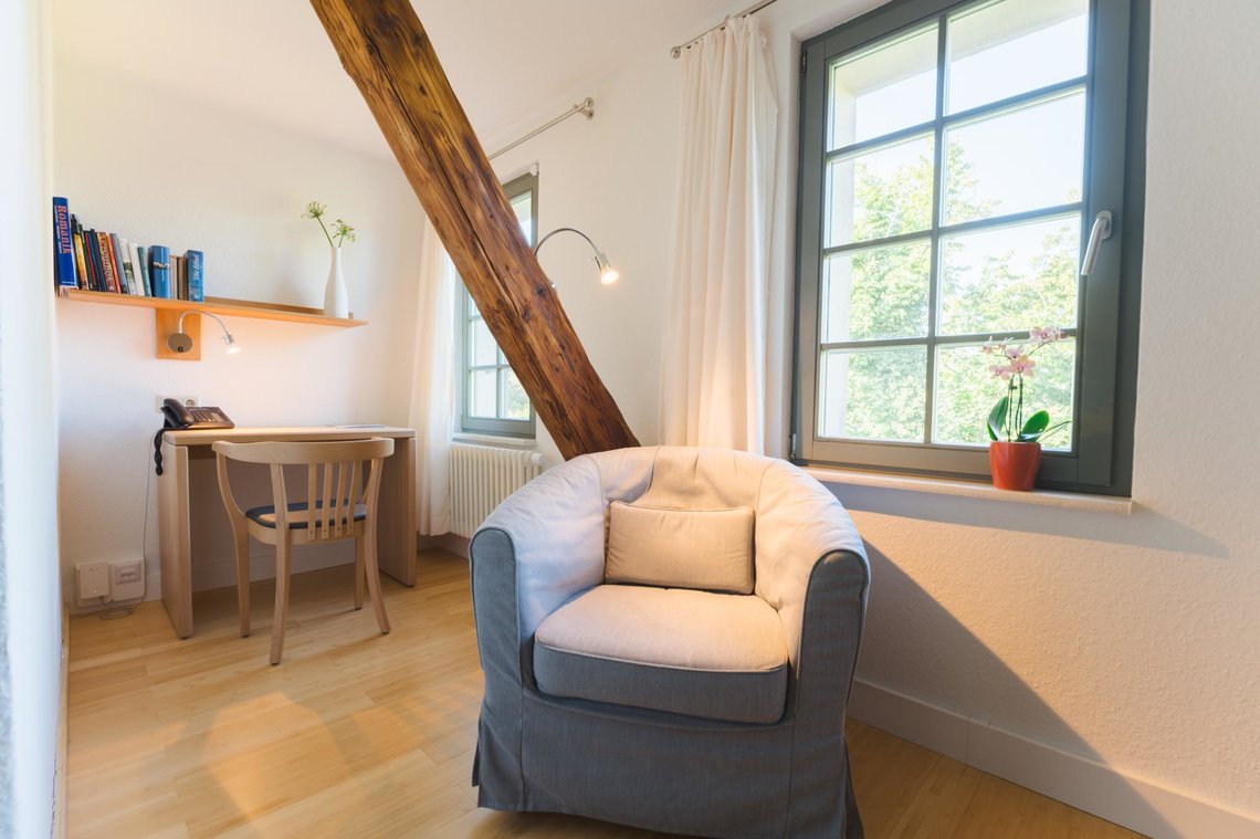 Ein grauer Sessel steht in einem Raum vor einem Schreibtisch mit Stuhl. Darüber hängt ein Bücherregal. Rechts vom Stuhl ist ein Fenster zu sehen.