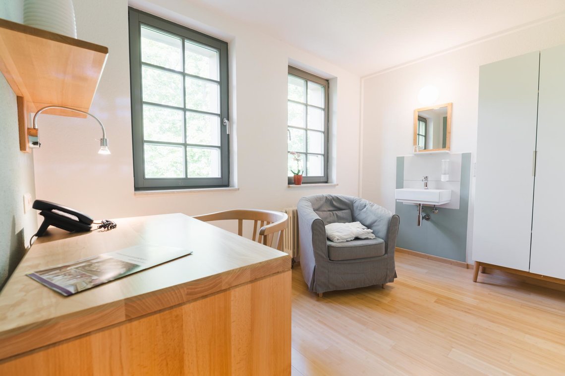 Ein Hotelzimmer  mit zwei Fenstern und davor einem grauen Sessel. Hinter dem Sessel ist ein Waschbecken mit Spiegel und rechts daneben ein weißer Schrank. Im Vordergrund links ist ein Schreibtisch mit einem Stuhl, einem Telefon und einer Lampe an der Wand. Über der Lampe ist ein Regalbrett.