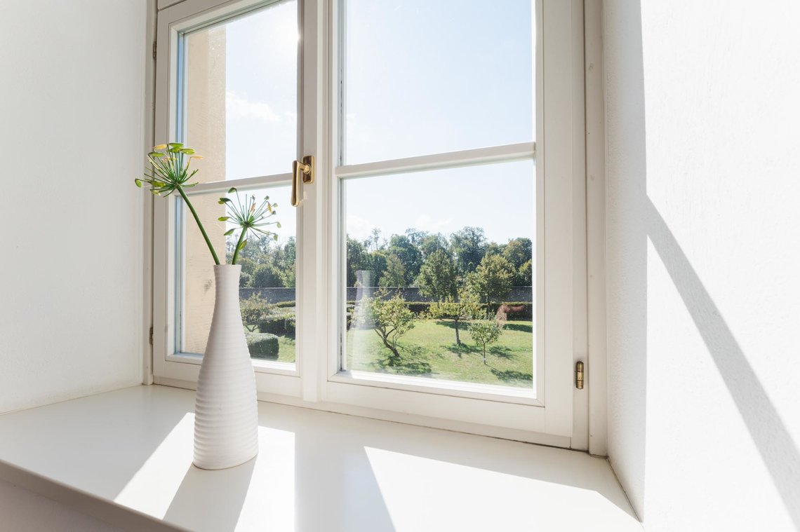 Zwei Blumen in einer weißen Vase vor dem Fenster, das aus dem barrierefreien Zimmer nach draußen zeigt. Zu sehen ist der Garten mit kleinen Bäumen