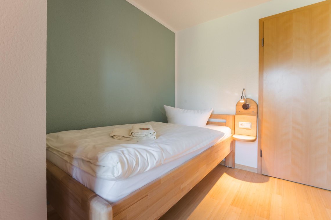 Ein Bett steht neben einer grünlichen Wand neben einer Tür. Auf dem Bett liegen zwei Handtücher und neben dem Bett ist eine Lampe mit einer Ablage.