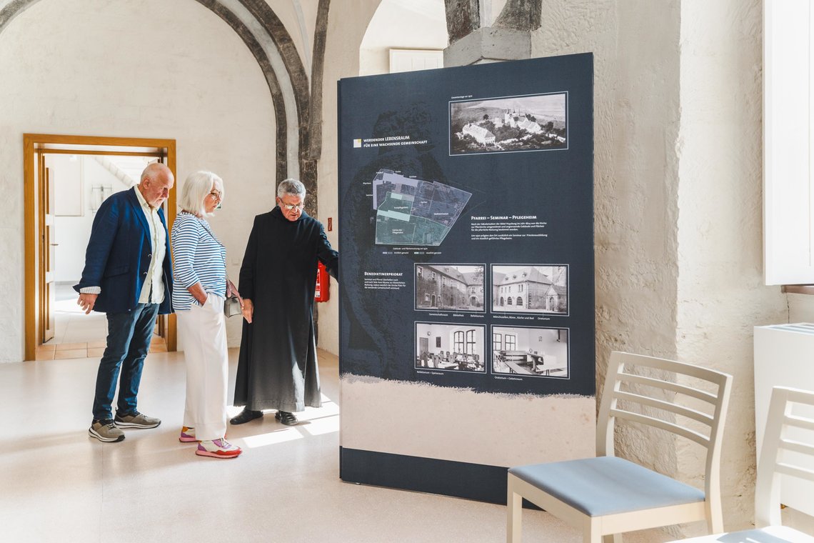 Ein Mönch und ein älteres Paar steht neben eine Informationstafel mit Texten und Bildern, die die Huysburg 1970 zeigen 