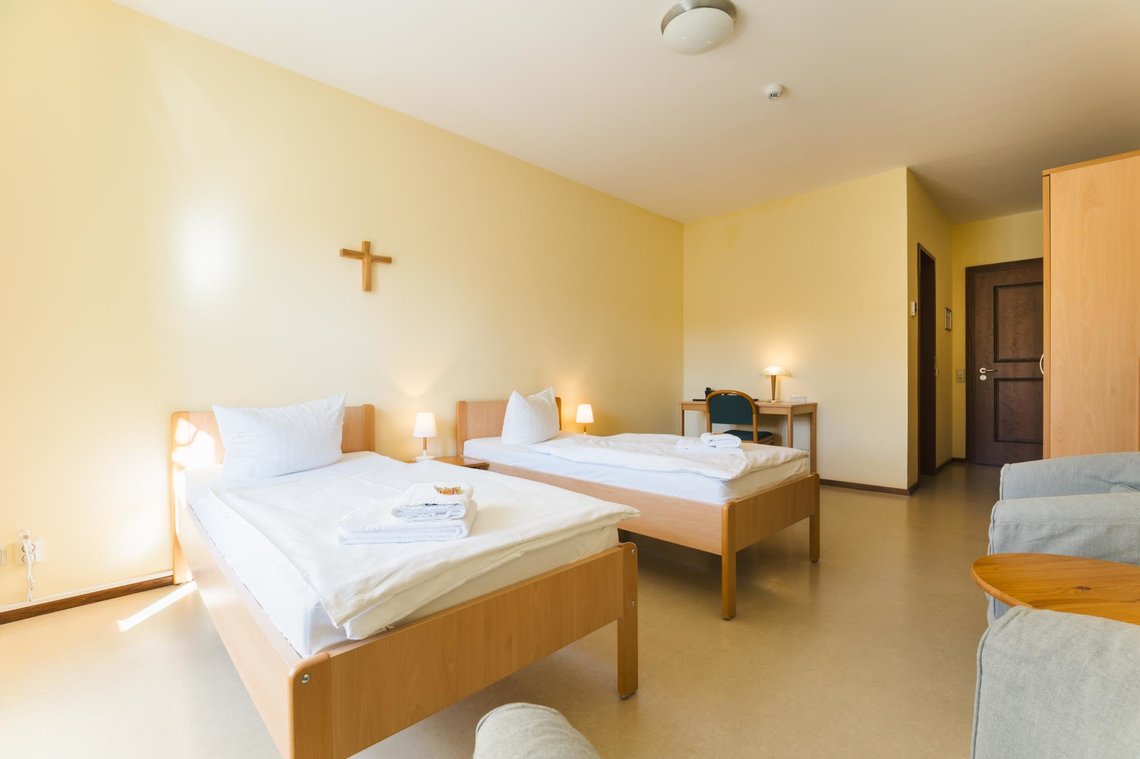 Ein helles, gelbes Zimmer mit zwei Einzelbetten mit zwei Nachttischen mit Lampe. Über den Betten hängt ein Kreuz. Rechts neben den Betten steht ein Schreibtisch mit einem Stuhl und einer Lampe. Daneben ist ein schmaler Flur mit 2 Türen. An der Rechten Wand steht ein Schrank, zwei graue Sessel und ein Beistelltisch.